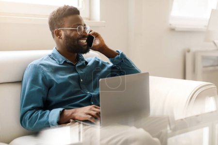 Photo for Happy black businessman in glasses calling by phone, working with laptop on couch in office interior, free space. Male entrepreneur talking with client - Royalty Free Image