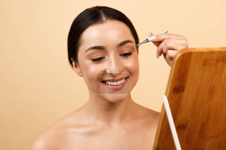 Téléchargez les photos : Young indian female plucking eyebrows with tweezers while looking in mirror, beautiful smiling hindu woman shaping her brows, enjoying self-care routine while standing over beige background - en image libre de droit