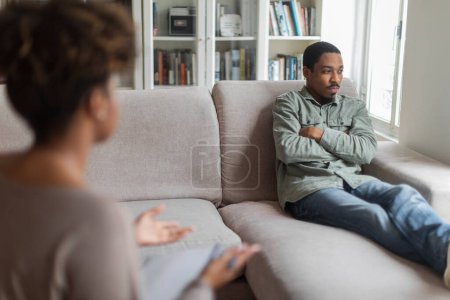 Foto de Reserved angry young black man resting on couch with arms crossed on chest, looking through the window at clinic, african american guy with addiction or PTSD attending psychotherapist woman - Imagen libre de derechos