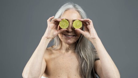 Téléchargez les photos : Vieille dame nue européenne joyeuse aux cheveux gris met des morceaux de citron vert à ses yeux comme des lunettes, s'amuse isolé sur fond gris, studio, panorama. Soins de beauté naturels, soins spa, publicité et offre - en image libre de droit