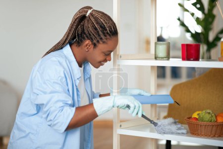Photo for Positive beautiful young african american woman in homewear cleaning dust at home, using feather duster, wearing rubber gloves, side view, copy space. Chores, house cleaning concept - Royalty Free Image