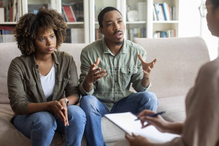 Foto de African american young husband and wife sitting on couch at family therapist office, fighting while visiting psychologist, emotional black man sharing his feeling with counselor, copy space - Imagen libre de derechos