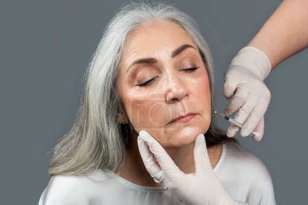 Photo for Calm old european lady with closed eyes getting beauty injection, enjoy anti-aging procedure with cosmetologist in gloves isolated on gray background, studio. Wrinkle treatment, skin care, cosmetology - Royalty Free Image