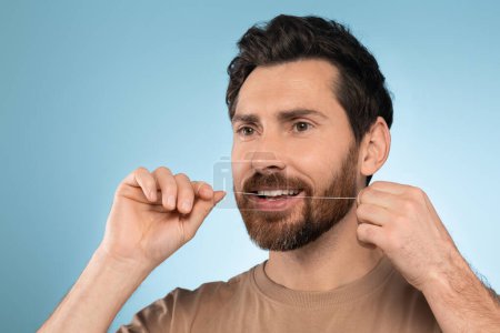 Foto de Retrato de hombre barbudo de mediana edad usando hilo dental y sonriendo, limpiando los dientes después de la comida, de pie sobre el fondo azul del estudio. Concepto de cuidado dental - Imagen libre de derechos