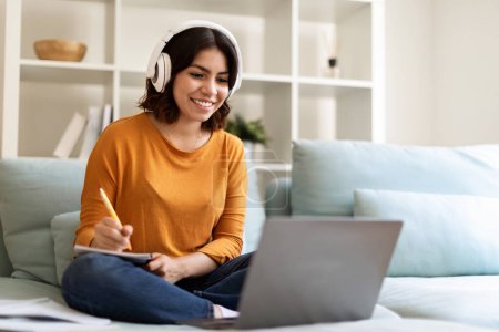 Foto de Young Arab Woman In Headset Study Online With Laptop From Home, Smiling Middle Eastern Female Watching Webinar On Computer And Taking Notes To Notepad, While Sitting On Couch In Living Room - Imagen libre de derechos