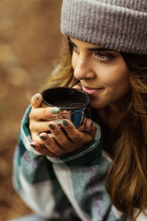 Foto de Smiling millennial caucasian woman in jacket and hat drink hot tea cup, travel in cold forest, enjoy adventure outdoor, vertical. Vacation, active lifestyle and camping, hikers sports and walking - Imagen libre de derechos