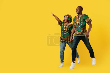 Photo for Excited happy black lovers in colorful african costumes holding hands, jumping in the air, pointing at copy space for advertisement and smiling, isolated on yellow studio background, full length - Royalty Free Image