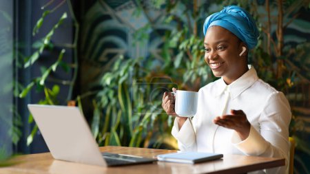 Téléchargez les photos : Amical jolie jeune femme noire en belle tenue entrepreneur indépendant travaillant à partir d'un café, assis à table, boire du café, en utilisant un ordinateur portable et des écouteurs, avoir une vidéoconférence, panorama, espace de copie - en image libre de droit