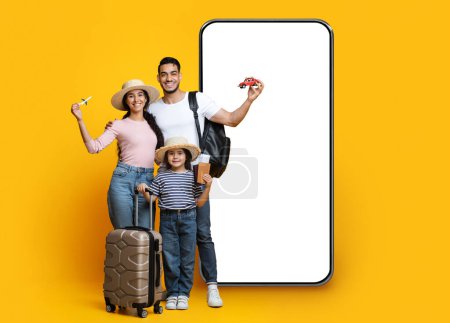 Foto de Family Travel Deal Concept. Happy middle eastern parents with little daughter holding plastic plane and car, posing by big phone with movkup on yellow background, mom, dad and child ready for vacation - Imagen libre de derechos
