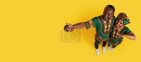 Photo for High angle view of cheerful positive loving black spouses wearing traditional african clothing embracing, pointing at copy space on yellow studio background, panorama - Royalty Free Image