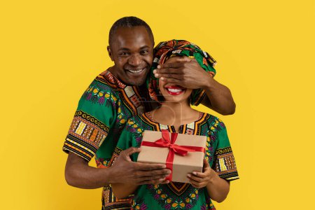 Photo for Loving handsome middle aged black man make surprise for his wife, closing excited lady eyes and holding gift box, couple wearing african outfits, yellow studio background, celebrating anniversary - Royalty Free Image
