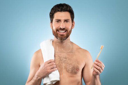 Photo for Portrait of handsome bearded man with towel on shoulder smiling at camera, standing with toothbrush over blue background. Dental care concept - Royalty Free Image