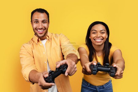 Téléchargez les photos : Happy young african american husband and wife with joystick have fun, playing computer online game together, isolated on yellow background, studio. Win, entertainment with modern gadget, ad and offer - en image libre de droit