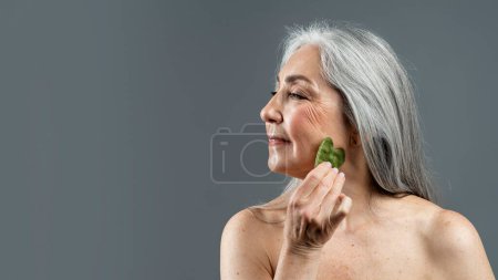 Téléchargez les photos : Heureuse vieille femme nue européenne heureuse avec des cheveux gris faisant massage du visage avec de la pierre de jade, isolée sur fond gris, panorama, studio. Soins de la peau et de beauté, soins spa, rajeunissement, publicité et offre - en image libre de droit
