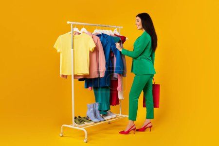 Photo for Young female shopaholic shopping and picking outfit, choosing colorful clothes on rail, standing over yellow background, full length. Fashion and trends - Royalty Free Image