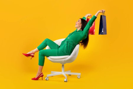 Téléchargez les photos : Joyeux shopaholic femme avec beaucoup de sacs à provisions colorés assis sur la chaise sur fond de studio jaune, espace de copie, vue latérale. Plan complet - en image libre de droit