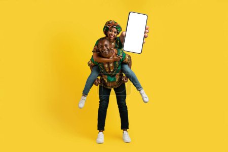 Photo for Loving african man and woman in traditional costumes having fun over yellow studio background, happy black lady piggybacking her husband, showing phone with white blank screen, mockup, copy space - Royalty Free Image