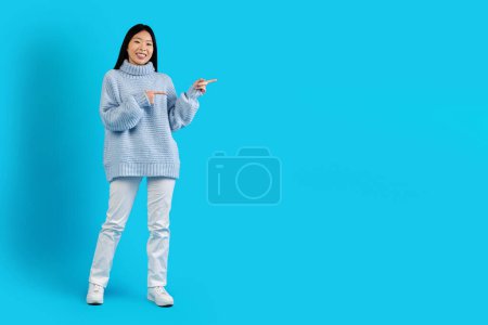 Téléchargez les photos : Mignon joyeux attrayant cheveux longs jeune femme asiatique en pull chaud et pantalon blanc posant sur fond bleu studio, pointant vers l'espace de copie pour l'annonce ou le texte, montrant belle offre, pleine longueur - en image libre de droit