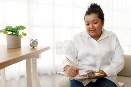 Photo for Chubby attractive young mixed race lady in formal outfit counselor working at office, sitting on couch, holding folder and pen, therapist taking notes while listening to patient request, copy space - Royalty Free Image