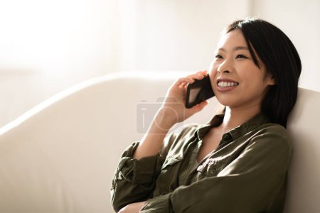 Téléchargez les photos : Happy cheerful smiling beautiful millennial asian woman in comfy casual outwear sitting on couch, having phone conversation with friend or lover at home, looking at copy space for ad, closeup - en image libre de droit