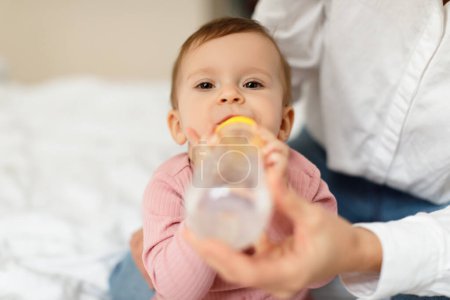 Foto de Mother giving her little daughter bottle of water feeding her sitting on bed at home, closeup shot, free space. Maternity leave, child care, parenting concept - Imagen libre de derechos