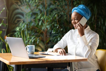Foto de Cheerful pretty young black woman business advisor working at coffee shop, female freelancer sitting at table alone, using modern computer, having phone conversation with client, copy space - Imagen libre de derechos