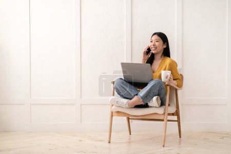 Téléchargez les photos : Joyeux heureux belle jeune femme asiatique dans occasionnel assis dans un fauteuil avec ordinateur portable, parler au téléphone, boire du café, regarder l'espace de copie et sourire. Travail à distance, concept freelance - en image libre de droit