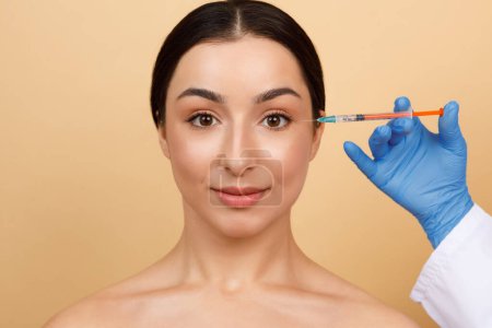Téléchargez les photos : Jeune femme indienne obtenant l'injection de Botox pour réduire les rides des yeux, médecin esthéticienne méconnaissable tenant la seringue près du visage féminin attrayant, prêt à faire une prise de vue, gros plan avec l'espace de copie - en image libre de droit