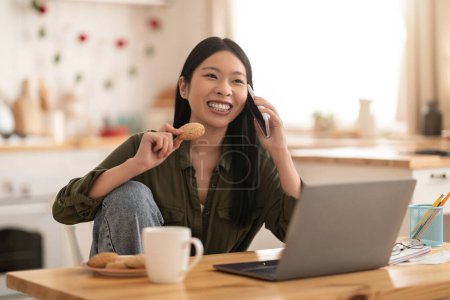 Téléchargez les photos : Cheerful happy beautiful young asian woman in casual sitting at kitchen with laptop, talking on phone, drinking coffee, eating cookies, looking at copy space and smiling, enjoying weekend at home - en image libre de droit