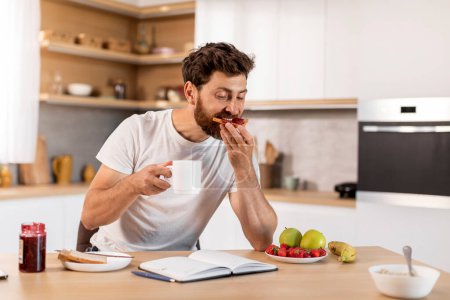 Foto de Busy handsome adult caucasian male in white t-shirt eats sandwich with jam, drinks coffee, reads book in modern kitchen interior. Breakfast, planning day and work, study, good morning, weekend home - Imagen libre de derechos