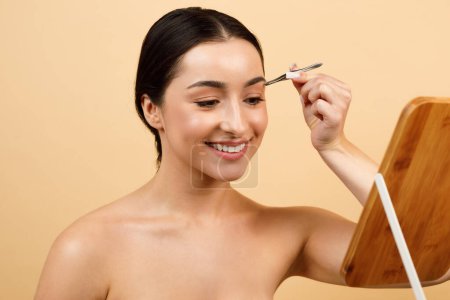 Téléchargez les photos : Portrait Of Young Indian Woman Standing Near Mirror And Plucking Her Eyebrows, Smiling Hindu Female Using Tweezers For Brows Correction While Standing Over Beige Studio Background, Free Space - en image libre de droit