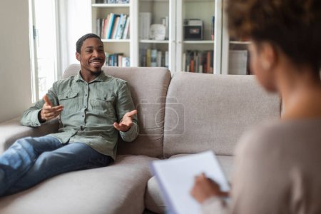 Téléchargez les photos : Inspired thrilled handsome young black man in casual reclining on couch at counselor office, sharing feelings, thoughts with therapist african american woman, enjoying therapy session. Mental health - en image libre de droit