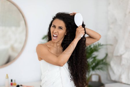 Téléchargez les photos : Angry sad millennial caucasian brunette lady in towel combing long curly hair, suffer from pain and tangled in bedroom interior, empty space. Routine procedures at home and problems with beauty care - en image libre de droit