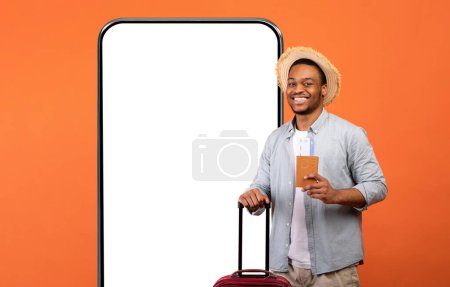 Photo for Young black man in casual clothes with suitcase, passport and tickets over big cell phone with empty screen, orange studio background, traveller booking plane flight online, using app, mockup, collage - Royalty Free Image