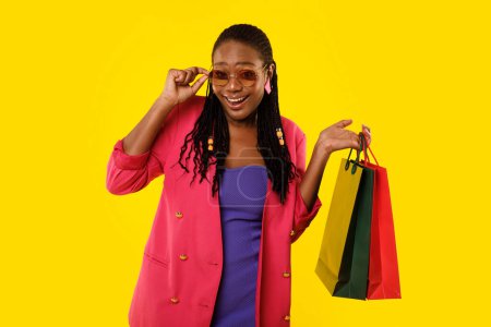 Foto de Oferta de compras. Feliz afroamericana Shopaholic Woman posando usando gafas de sol y sosteniendo bolsas de compras sonriendo a la cámara en el fondo amarillo del estudio. Gran concepto de ventas estacionales - Imagen libre de derechos