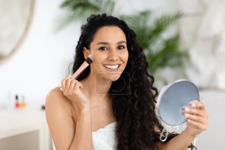 Téléchargez les photos : Glad millennial caucasian brunette lady with long hair in towel uses makeup brush, looks in mirror in white bedroom interior. Nude makeup routine at home and good morning, beauty care with cosmetics - en image libre de droit