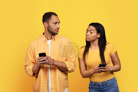 Photo for Curious young african american wife looking at husband smartphone, isolated on yellow background, studio. Jealousy, problems in relationship and love, gadget addiction, social network, ad and offer - Royalty Free Image