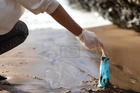 Téléchargez les photos : Female volunteer picking up used disposable medical face masks on the beach, cleaning coastal zone, closeup. The environmental problem after the coronavirus pandemic - en image libre de droit