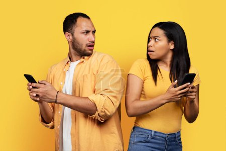 Téléchargez les photos : Shocked frightened young african american man and woman with smartphones looking at each other, isolated on yellow background, studio. Relationship problems, jealousy and gadget addiction, bad news - en image libre de droit