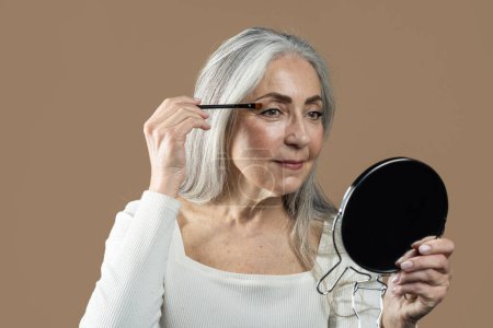 Photo for Smiling senior european female with gray hair applies mascara with brush on eyelashes, looks in mirror, isolated on brown background. Nude makeup, anti-aging cosmetics, beauty care, ad and offer - Royalty Free Image