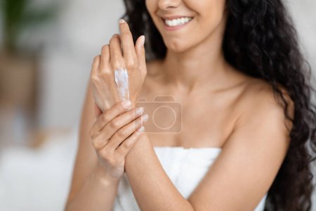 Photo for Glad millennial caucasian brunette lady with long hair in towel applies cream on hands in minimalist bedroom interior, enjoy free time cropped, close up. Skin care, routine at home and good morning - Royalty Free Image