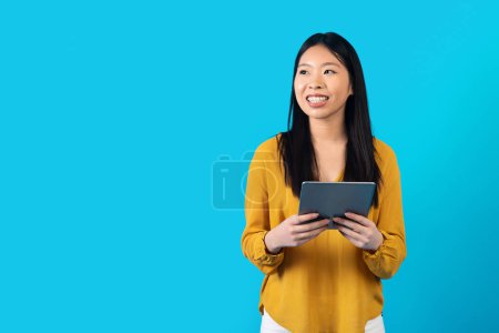 Foto de Alegre sonriente atractiva joven asiática utilizando almohadilla digital sobre fondo de estudio azul, mirando espacio de copia y sonriendo, comprobando más reciente aplicación móvil, compras, banca, estudiar en línea - Imagen libre de derechos