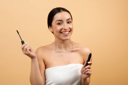 Téléchargez les photos : Beautiful Young Indian Woman Holding Mascara And Smiling At Camera, Happy Hindu Lady Wrapped In Towel After Bath Applying Makeup, Getting Ready While Standing Over Beige Studio Background, Copy Space - en image libre de droit