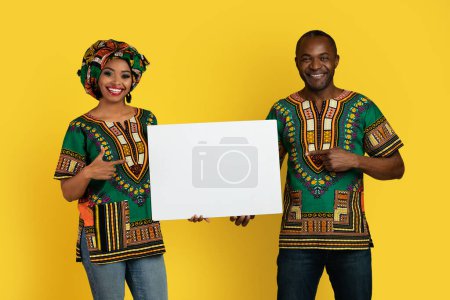 Téléchargez les photos : Emotional cheerful happy beautiful black couple in colorful traditional african costumes showing white blank placard with mockup for advertisement or text, yellow studio background - en image libre de droit