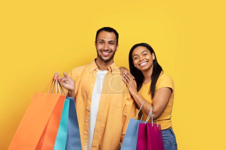 Téléchargez les photos : Glad young african american male and woman shopaholics hold many packages with purchases, isolated on yellow background, studio. Shopping together, sale and buyers enjoy buying and people emotions - en image libre de droit
