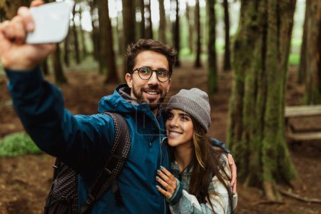Téléchargez les photos : Joyeux homme caucasien millénaire embrasser dame en veste et sac à dos, prendre selfie au téléphone dans la forêt froide, profiter de l'aventure ensemble en plein air. Photo pour blog, vacances actives et camping, randonneurs à pied - en image libre de droit