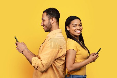 Photo for Smiling young african american family back to back, chatting on smartphones, isolated on yellow background, studio. Relationship and love, app for surfing, addiction to gadget, social network and blog - Royalty Free Image