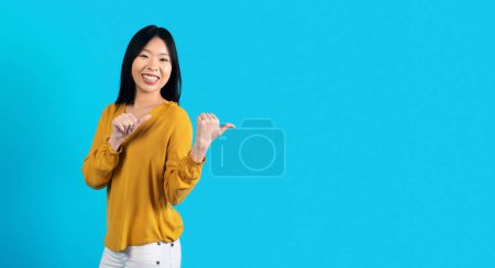 Téléchargez les photos : Studio plan de joyeuse belle dame millénaire asiatique en belle tenue posant sur fond bleu, pointant vers l'espace de copie et souriant, jeune femme chinoise montrant belle offre, panorama - en image libre de droit