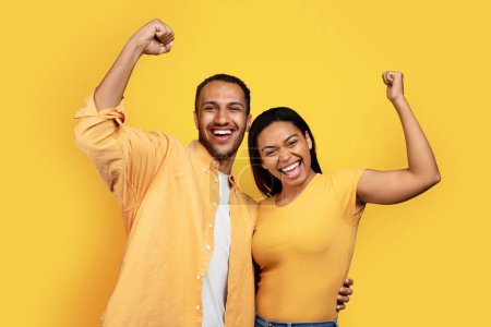 Téléchargez les photos : Cheerful young african american guy and woman hug, raise hands and make victory and success gesture, isolated on yellow background, studio, free space. Win emotion and sign, lifestyle, ad and offer - en image libre de droit