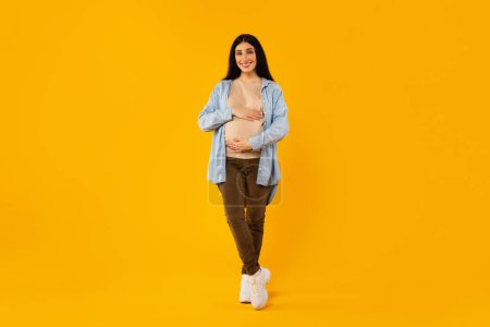 Photo for Full length shot of happy pregnant woman touching belly and smiling, posing over yellow studio background, smiling expectant lady enjoying healthy pregnancy time - Royalty Free Image